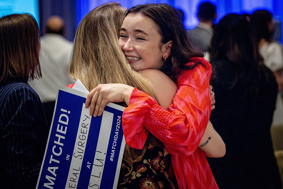 Two medical students hug. One is seen from the back, the other is seen smiling broadly, holding a sign that says "I Matched! In General Surgery and SLU!
